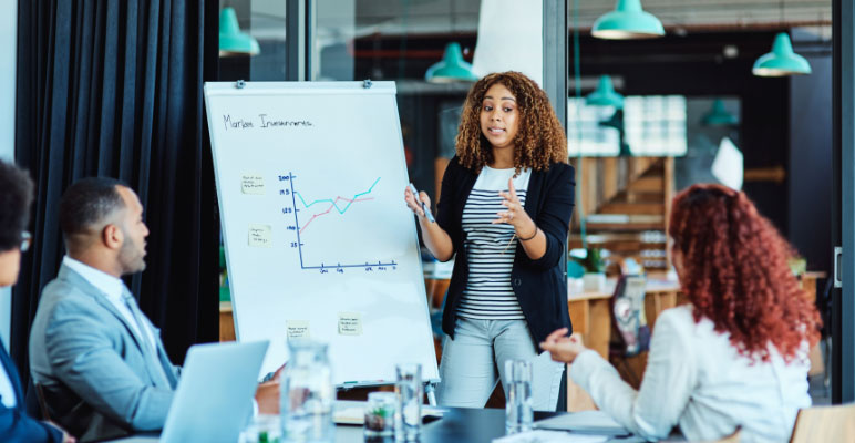 Photograph of man standing in front of three people sitting at desks with laptops while he is pointing at a bar graph on a large screen.